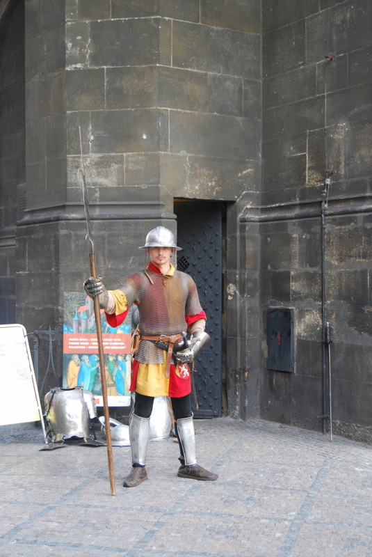 6 - A person dressed in a replica medieval suit of armor and holding a polearm, standing on a cobblestone street in front of a historic stone building in Prague